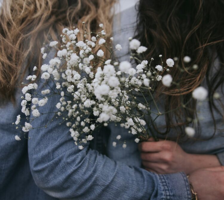 Two women with their arms around each other's backs, holding white flowers
