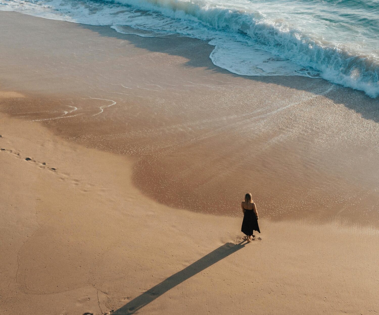 A view of a beach from above, with a women standing at the edge of the waves watching the water