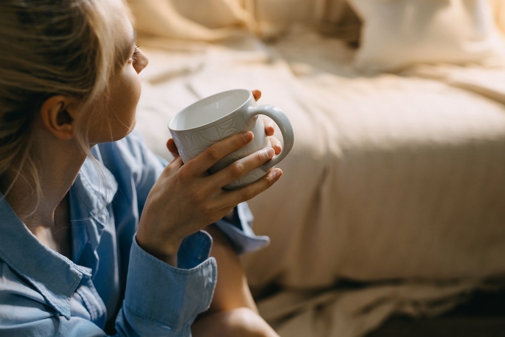 A woman taking a moment to herself with a cup of coffee beside an unmade bed
