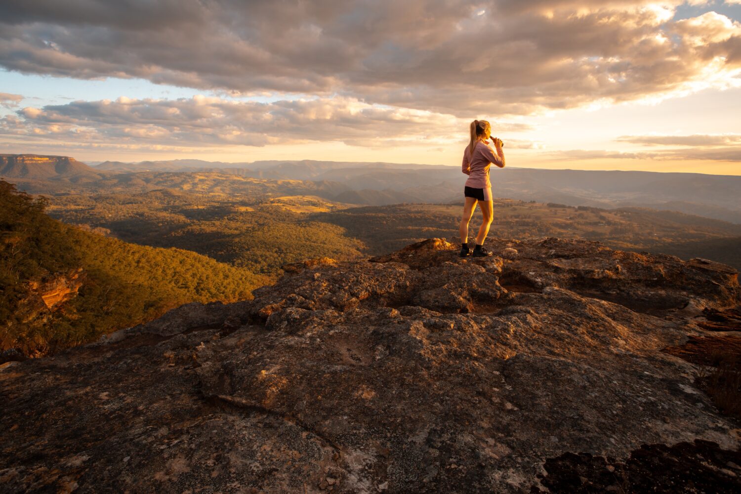 A woman standing on top of a lookout in the Blue Mountains, looking at the view and drinking from a drink bottle