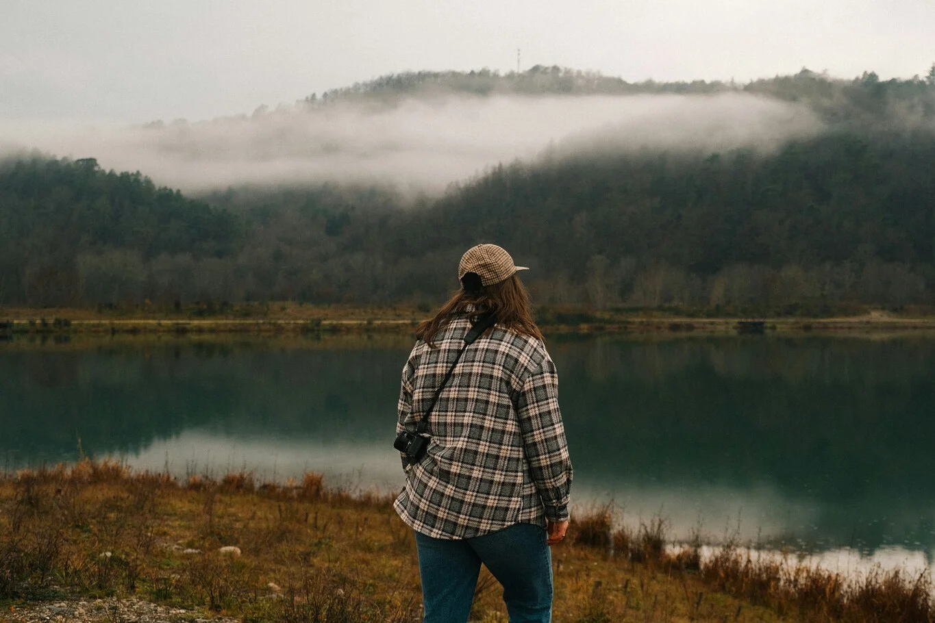 Person standing beside a lake looking toward a forested hillside.