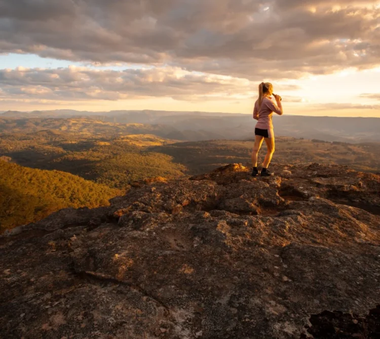 A woman standing on top of a lookout in the Blue Mountains, looking at the view and drinking from a drink bottle