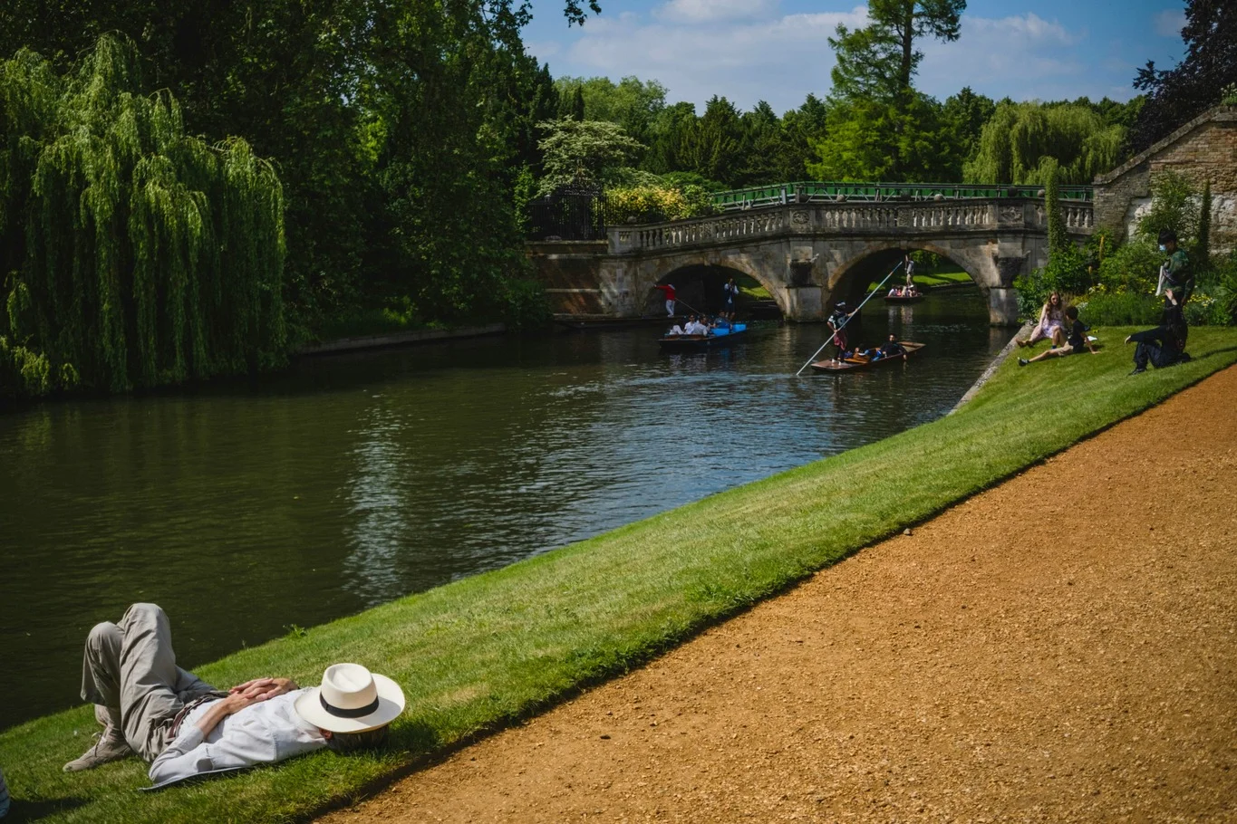 Person lying on the grass beside a river in a park with trees, a bridge, and people on boats.