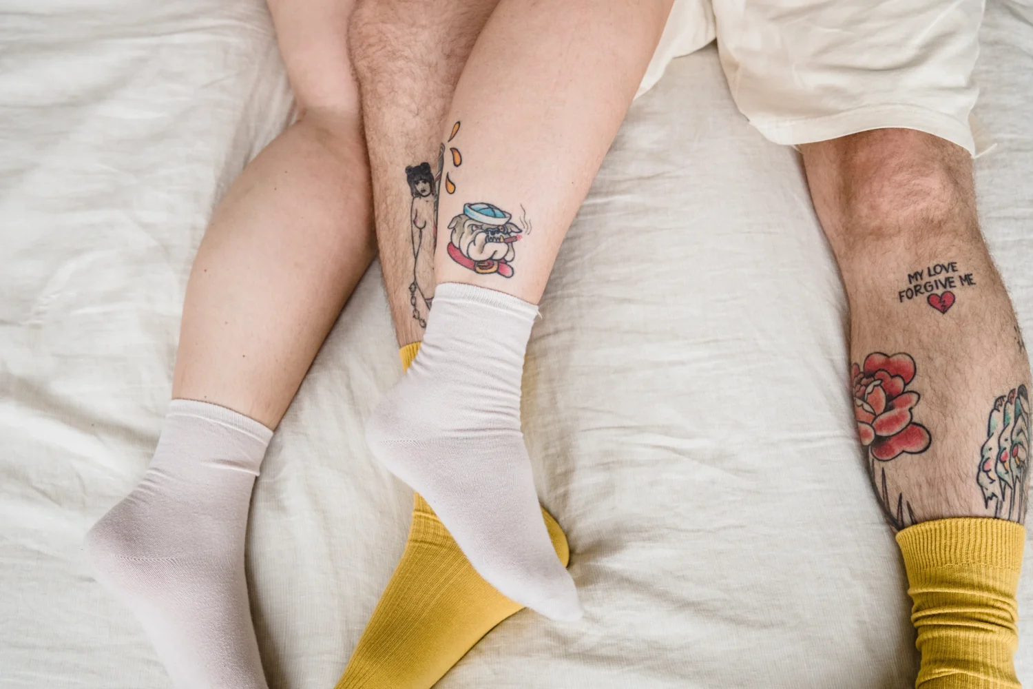Two people laying on a bed with intertwined legs, mismatched socks, and visible tattoos, creating a casual and intimate atmosphere.