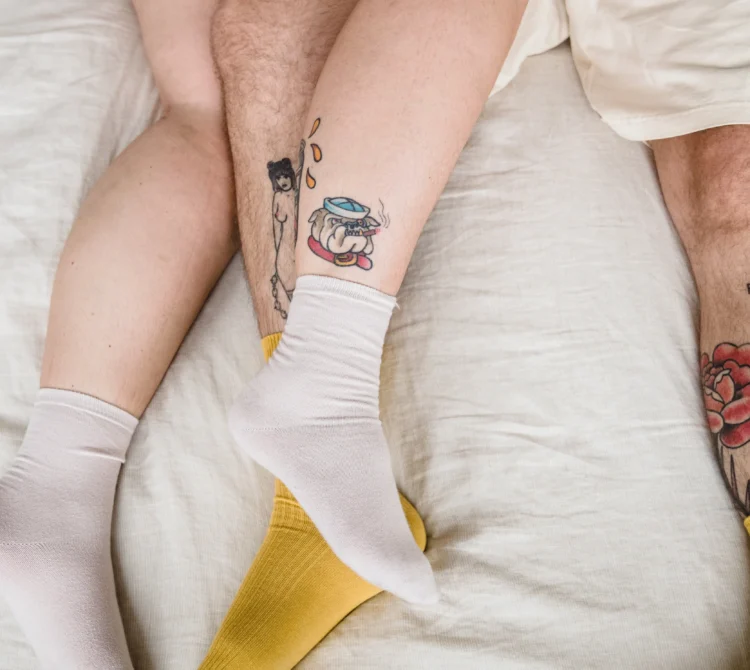 Two people laying on a bed with intertwined legs, mismatched socks, and visible tattoos, creating a casual and intimate atmosphere.