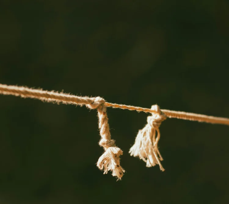 Close-up of two frayed rope tassels tied onto a thinner horizontal cord, softly lit against a dark blurred background.