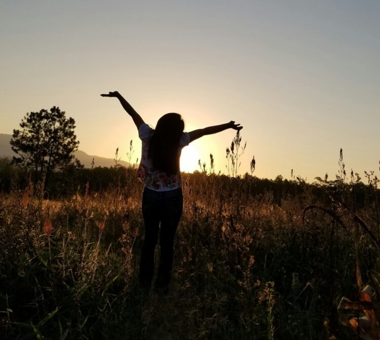 A woman standing in a field at sunset with arms upraised joyfully