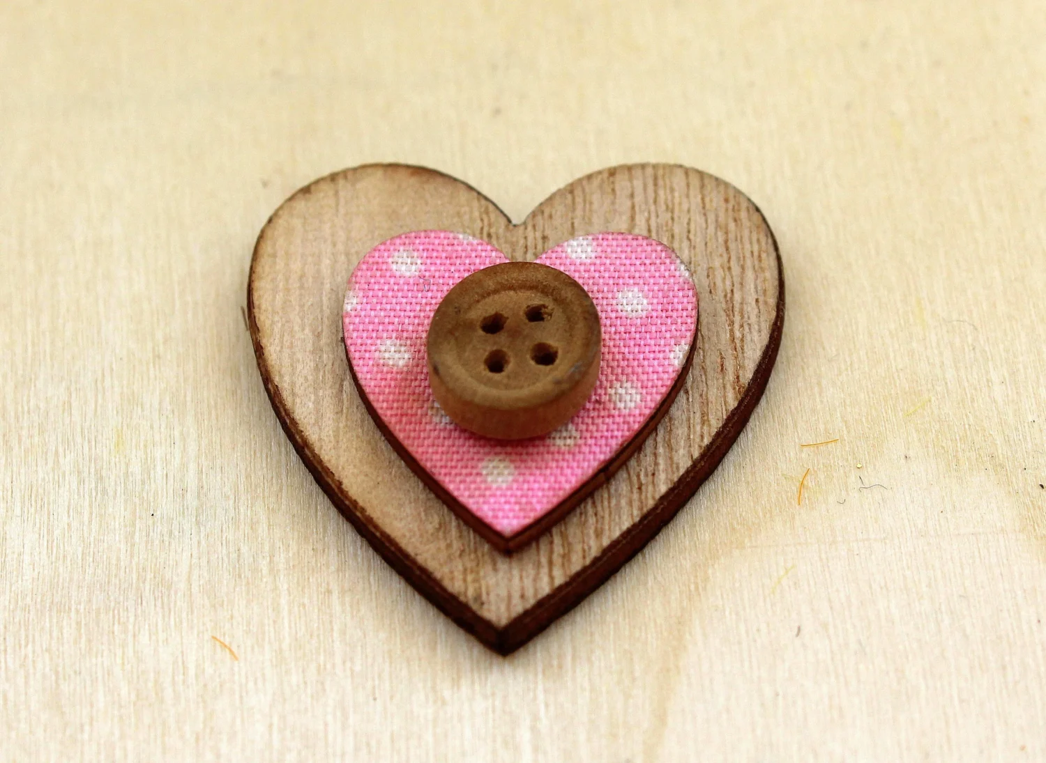 A small decorative heart made from layered wood and fabric, sits on a light wooden surface.