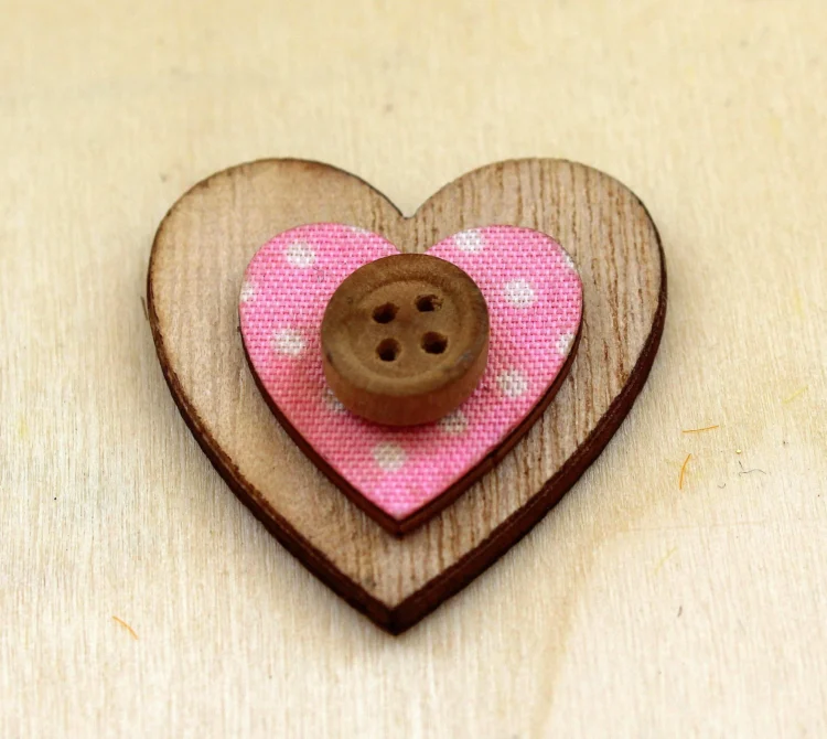 A small decorative heart made from layered wood and fabric, sits on a light wooden surface.